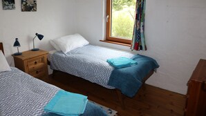 A bedroom with two beds, a window with curtains, a wooden nightstand, and a wall with three pictures. - The Song House (Falcarragh)