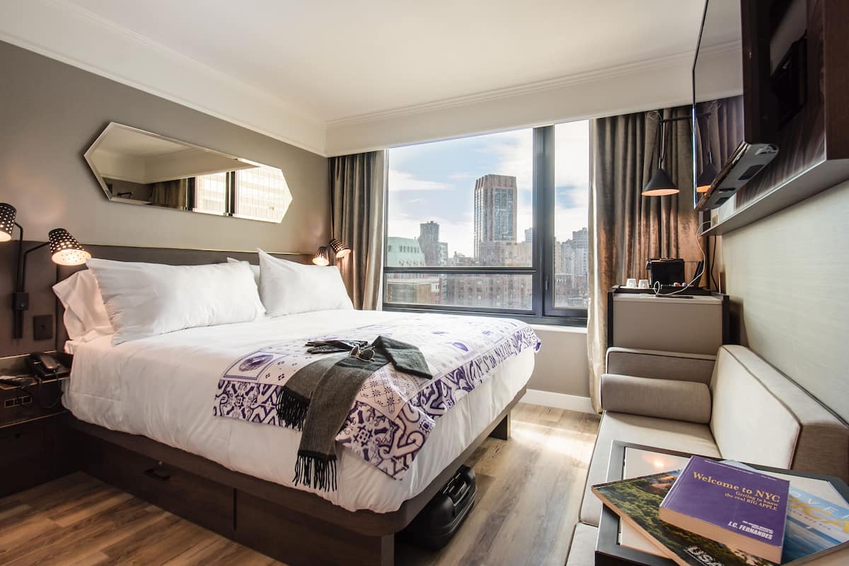 Ein modernes Hotelzimmer mit einem großen Bett, Blick auf die Stadt, einer Couch und einem Nachttisch mit Lampe.