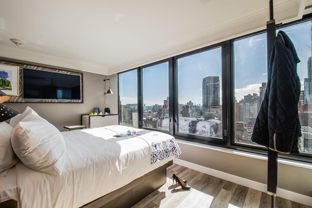 Ein modernes Schlafzimmer mit einem großen Bett, einem an der Wand montierten Fernseher und Blick auf die Stadt durch große Fenster.