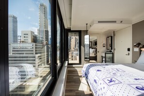 Ein modernes Hotelzimmer mit großem Fenster und Blick auf die Stadt, einem ordentlich bezogenen Bett und einem begehbaren Kleiderschrank.