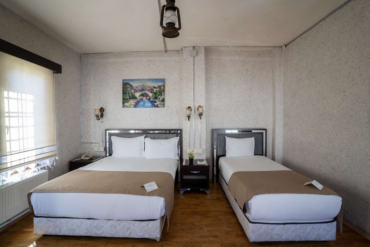 Habitación de hotel con dos camas, un cuadro en la pared, una ventana y una mesita de noche con una lámpara.