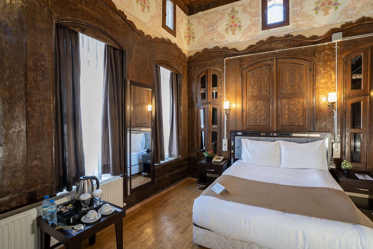 Una habitación de hotel con una cama grande, paneles de madera, una mesita con una tetera y una mesita de noche con una lámpara.