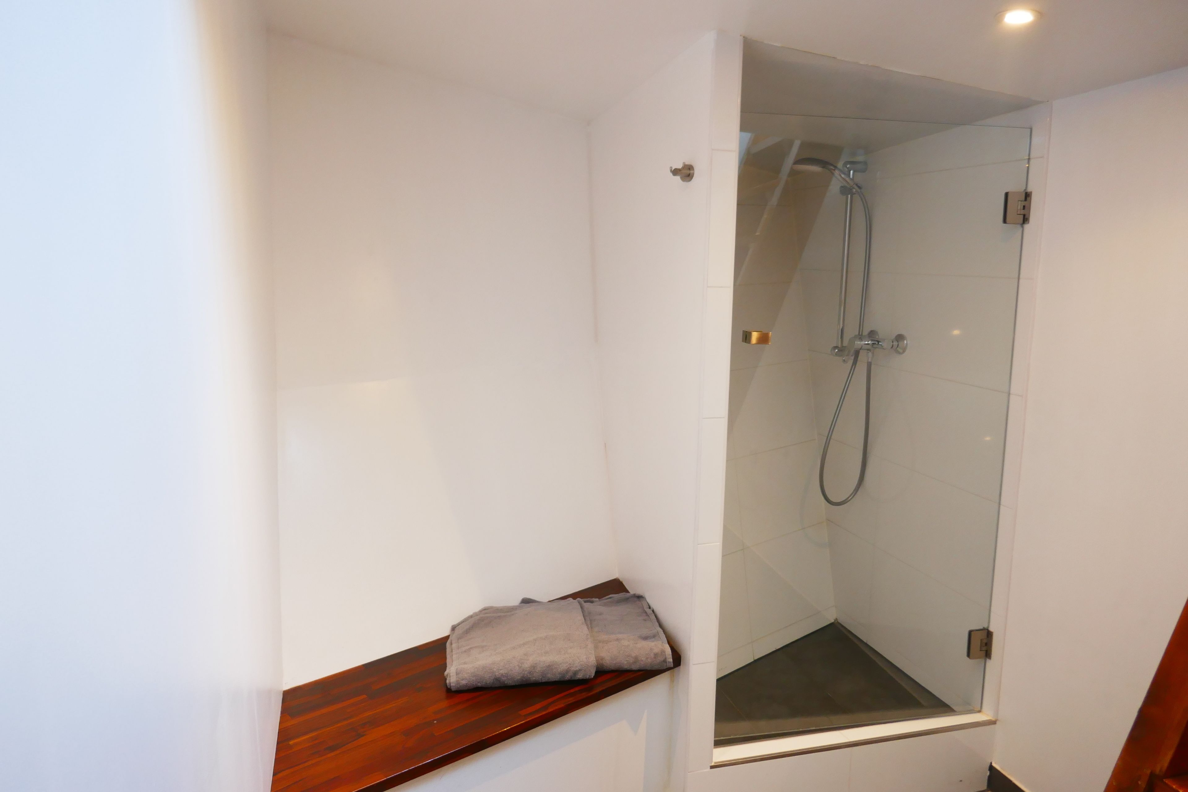Une petite salle de bain moderne avec une douche à parois de verre, une étagère en bois sur laquelle est posée une serviette, et des murs blancs.