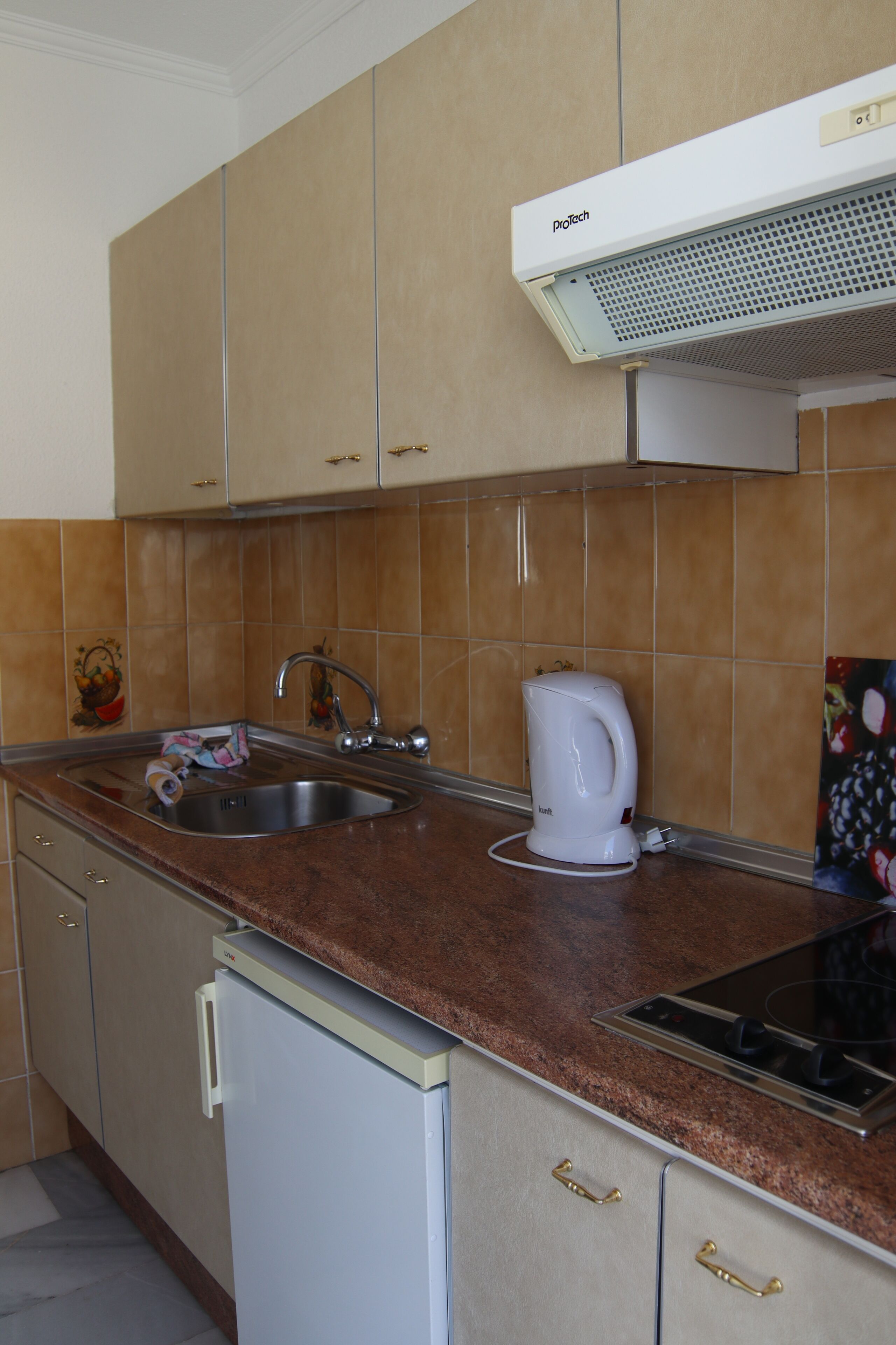 Una cocina con armarios color beige, un lavamanos de acero inoxidable, una tetera blanca y una estufa integrada.