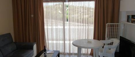 A living room with a sofa, a coffee table, a dining table with chairs, and a view through a glass door.