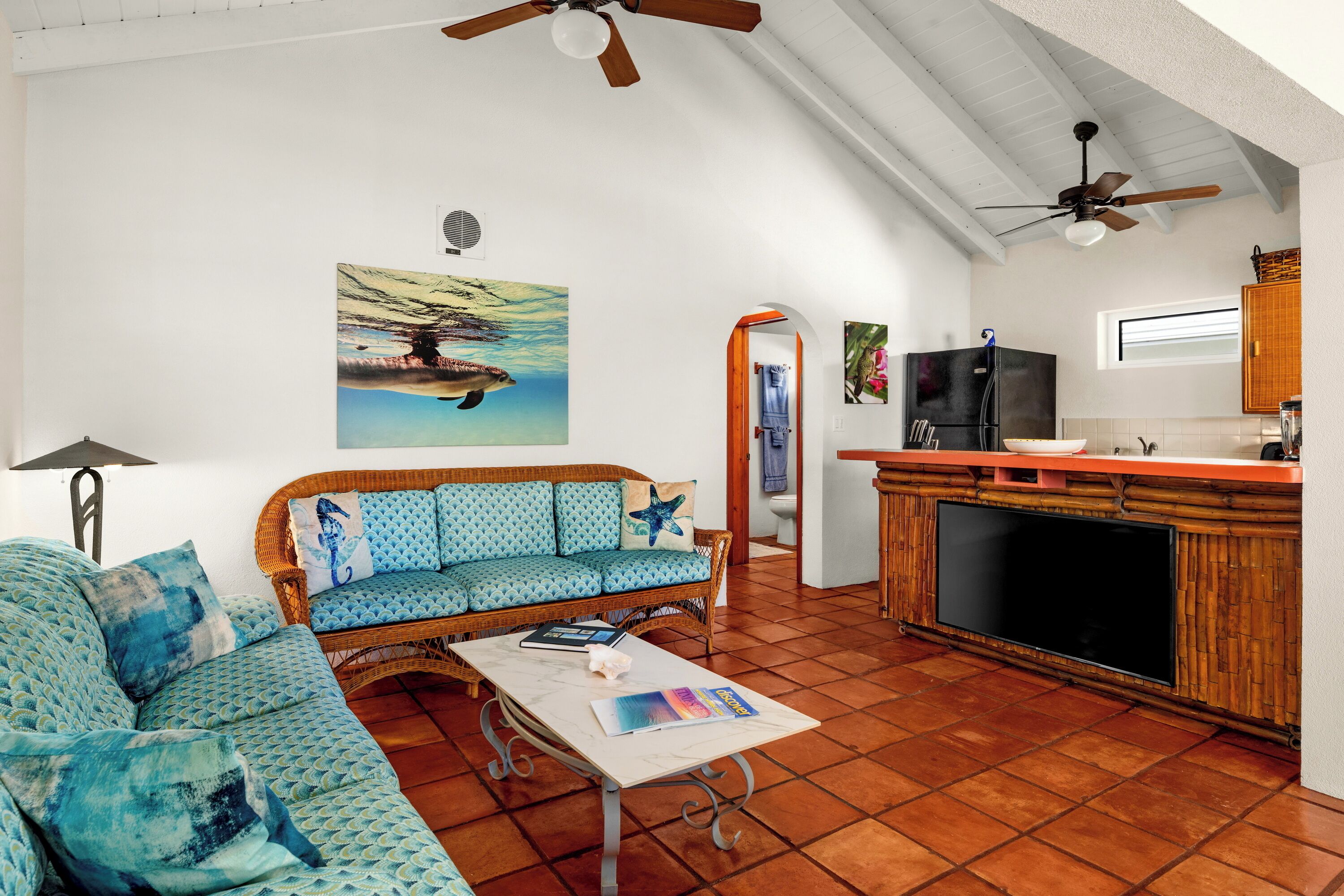 A living room with a sofa, coffee table, and a television. There is a kitchen area with a refrigerator and a window.