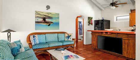 A living room with a sofa, coffee table, and a television. There is a kitchen area with a refrigerator and a window.