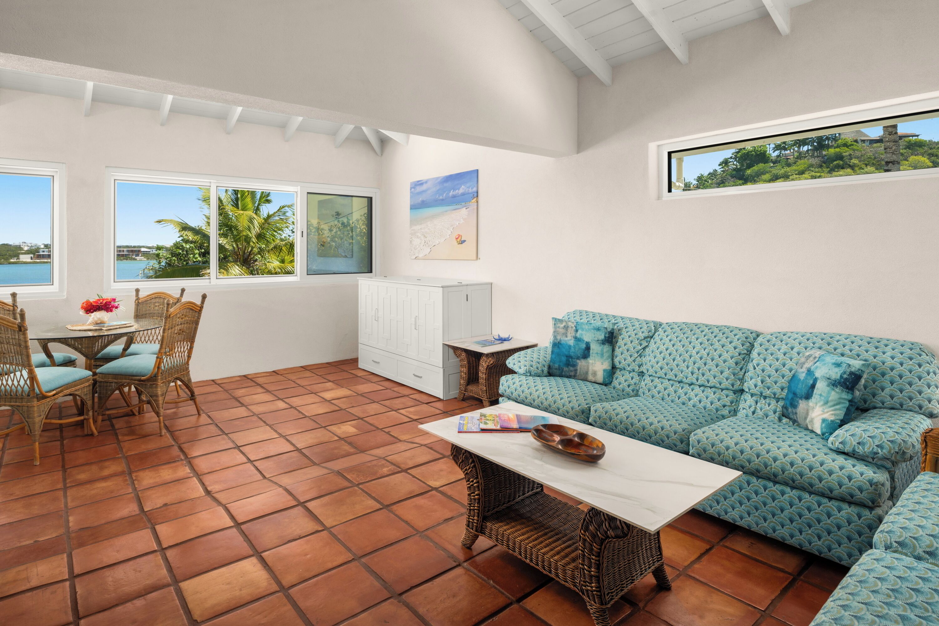 A spacious living area with a tiled floor, a sofa, a coffee table, a dining table with chairs, and large windows offering a view of the sea.