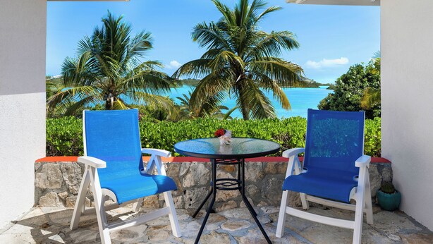 Un patio con dos sillas azules y una mesa pequeña, con vistas a una playa tropical con palmeras y el mar.