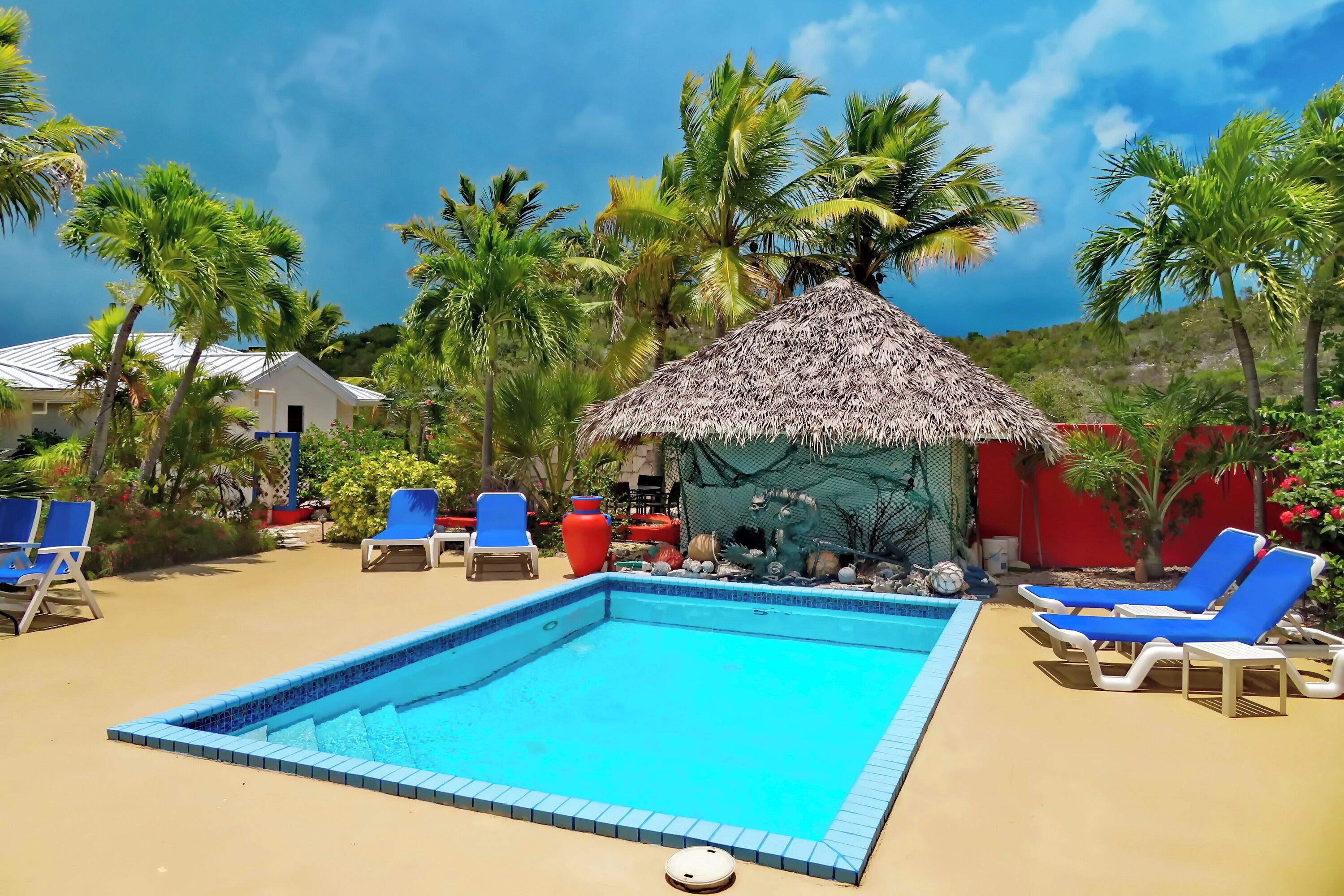 A swimming pool with blue tiles, surrounded by blue lounge chairs and a thatched-roof structure.