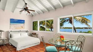 Un dormitorio con cama, ventilador de techo, ventana con vista a palmeras y agua, y una zona de comedor con mesa y sillas.