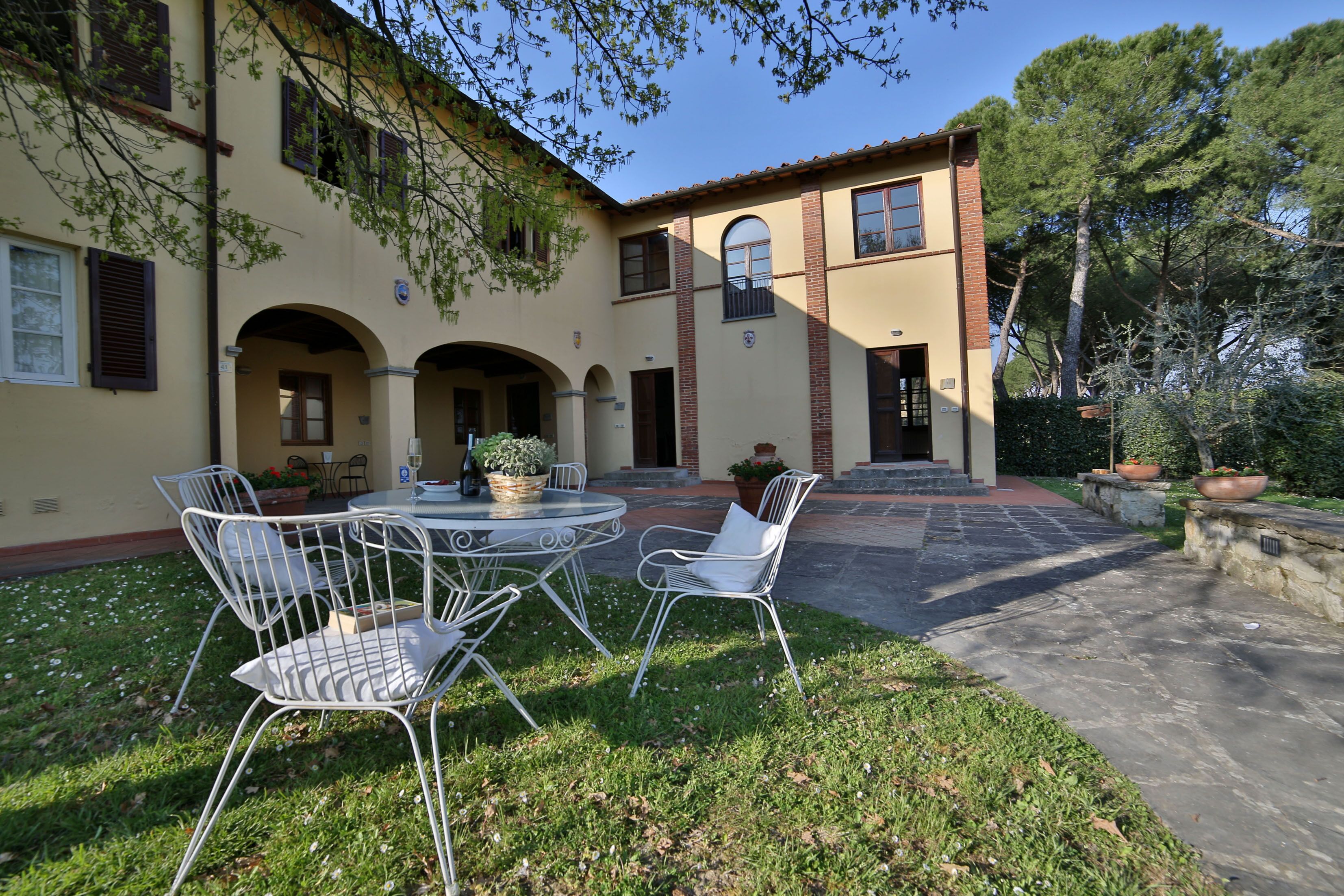 A courtyard with a round table and four white metal chairs, surrounded by a well-maintained lawn and potted plants.