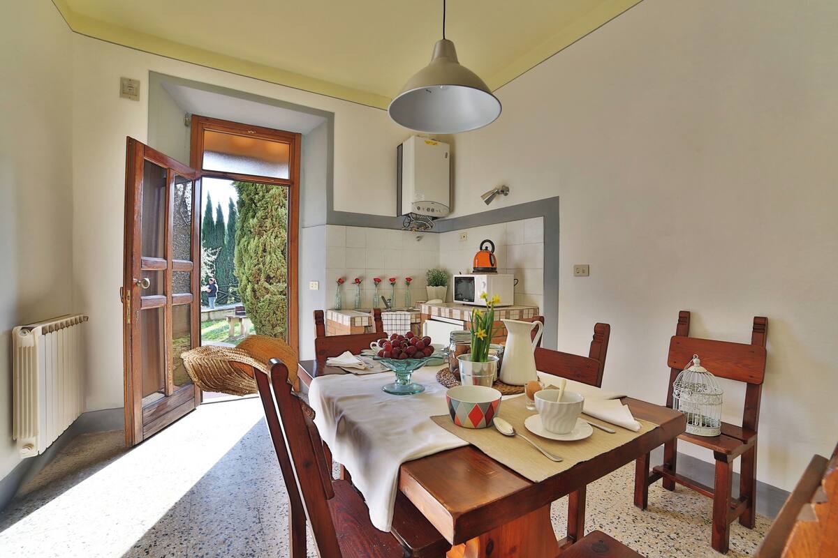 Un coin repas avec une table dressée pour deux personnes, un radiateur, une fenêtre donnant sur un espace verdoyant et un radiateur fixé au plafond.
