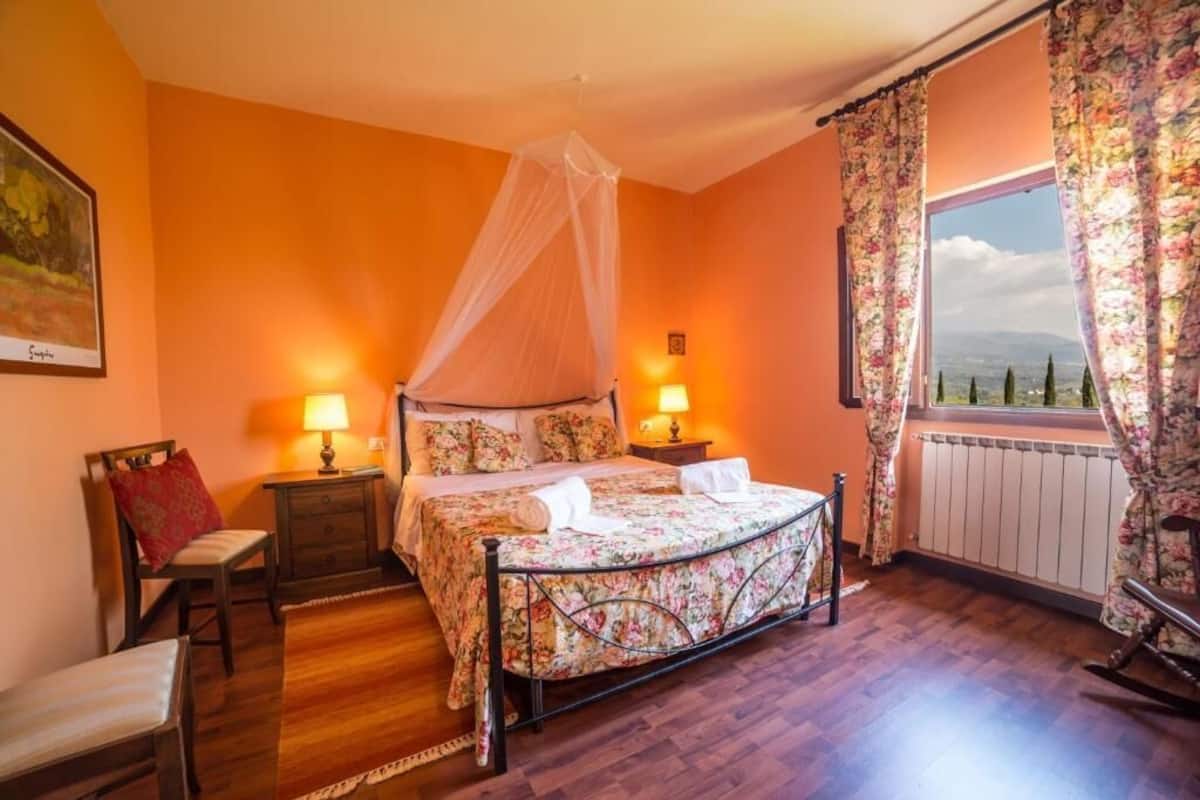 Une chambre à coucher avec un lit, des tables de chevet, une chaise, une fenêtre avec des rideaux et une vue.
