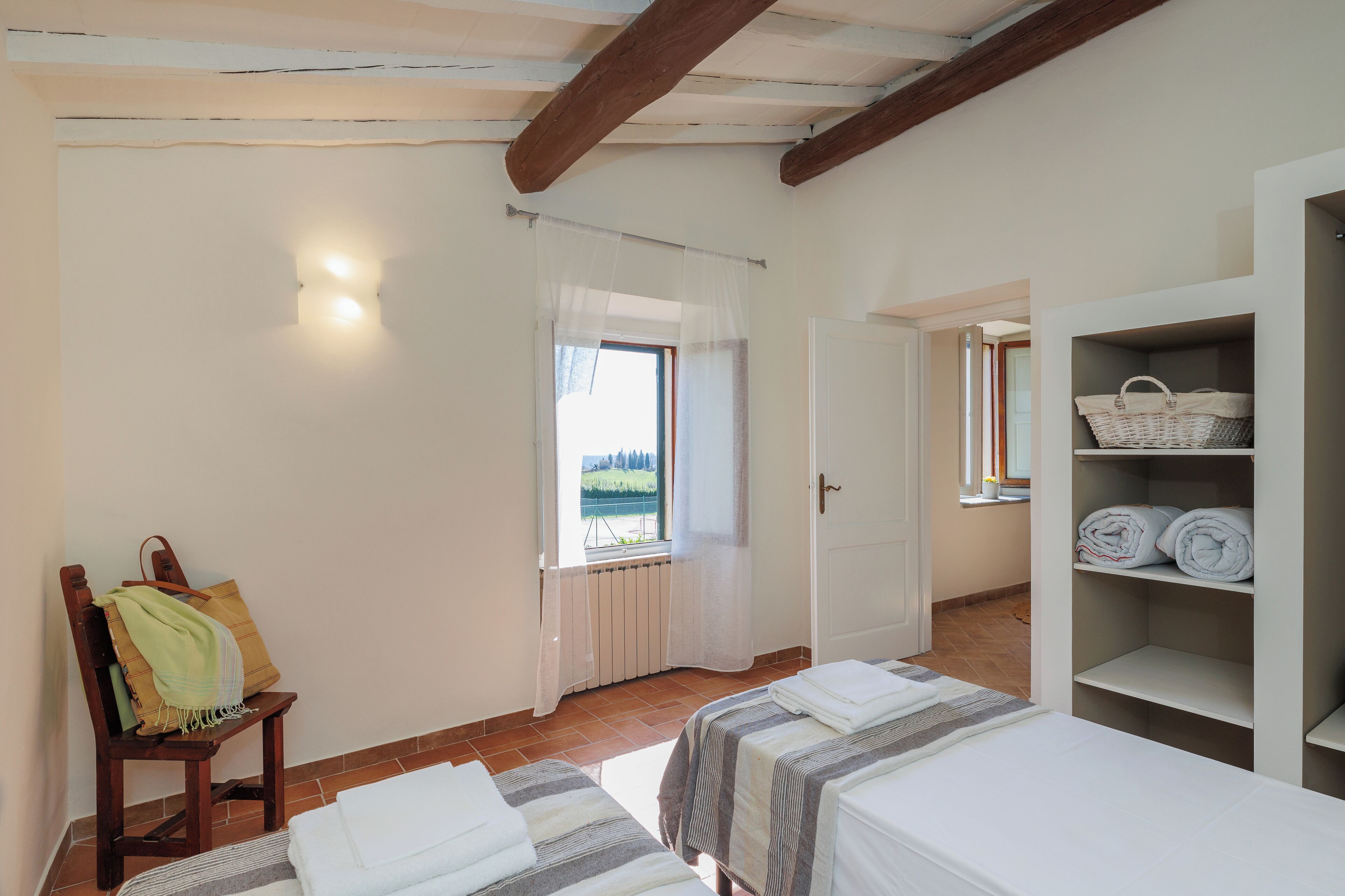 A room with two beds, a chair with a blanket, a shelf with towels, and a window with a view.