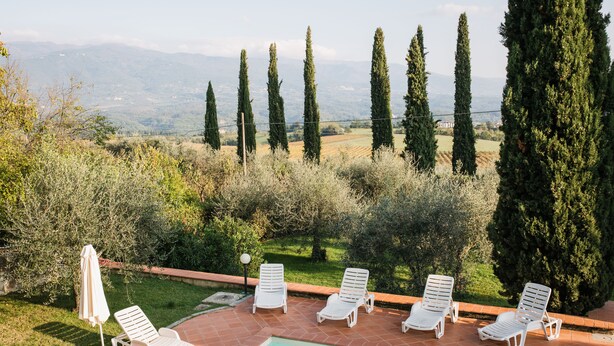 Un espace aménagé au bord de la piscine, avec des chaises longues et un parasol, entouré de verdure et de grands arbres, offrant une vue sur un paysage vallonné.