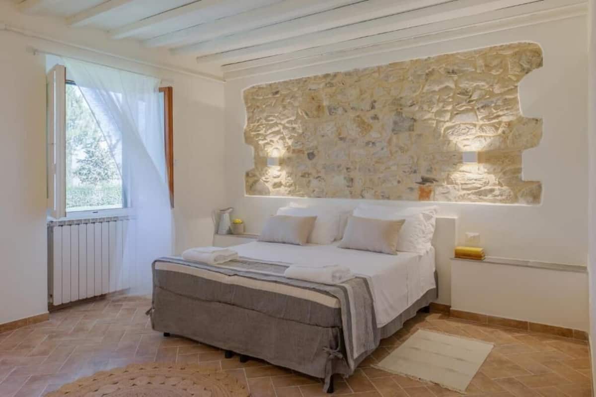Une chambre avec un mur en pierre, un lit avec une literie blanche, un radiateur et une fenêtre avec des rideaux.