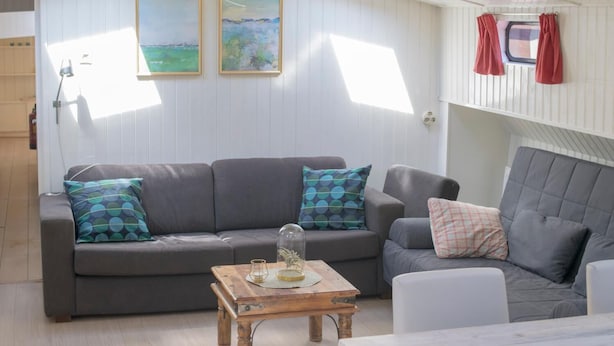 A cozy living room with a grey sofa, a wooden coffee table, and a white armchair. There are two framed paintings on the wall and a window with curtains.