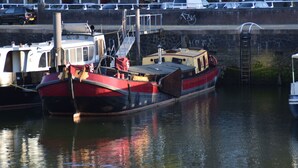 A red and black boat docked at a marina with a stone wall and parked cars in the background.