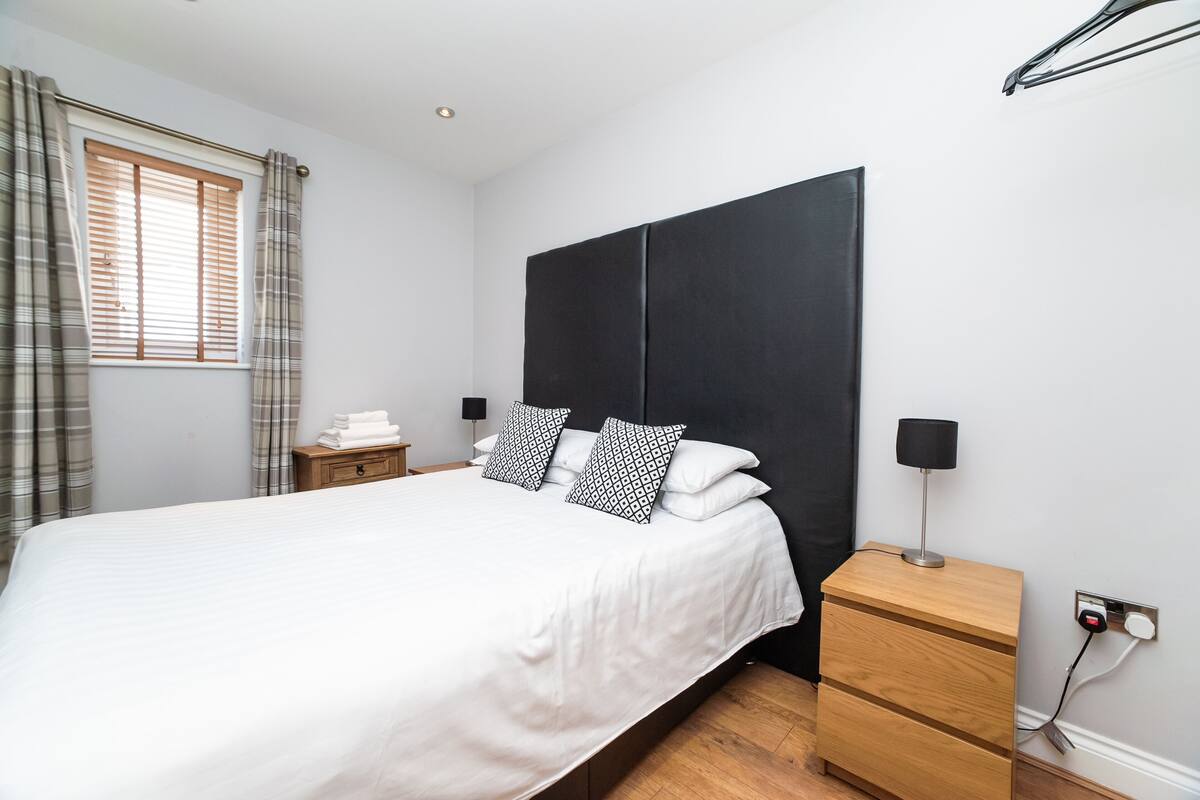 A neatly made bed with white bedding and black and white patterned pillows, a bedside table with a lamp, and a window with curtains.