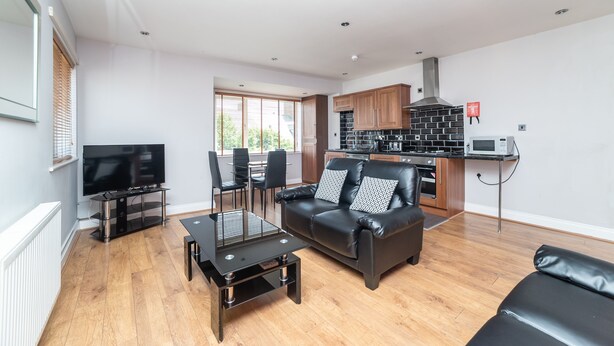 A modern living room with a black leather sofa, a glass coffee table, and a flat-screen TV. A kitchen with wooden cabinets and a microwave is visible in the background. - The New Mill Apartments (Newcastle-upon-Tyne)