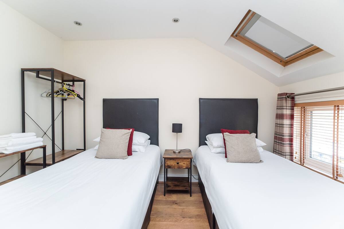 A hotel room with two beds, a small table, a wardrobe, and a skylight.