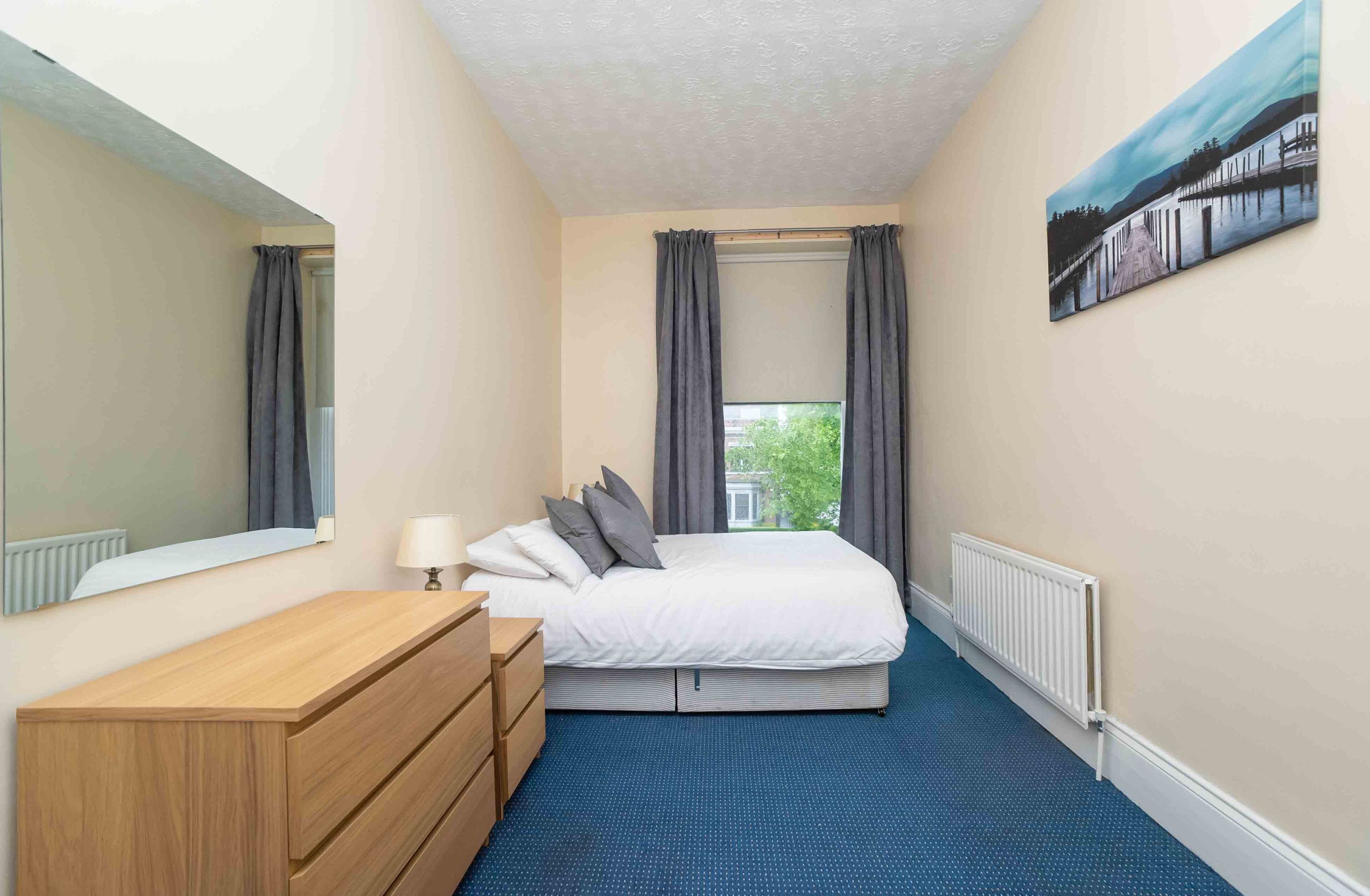 A bedroom with a bed, a dresser, a mirror, a window with curtains, a radiator, and a painting on the wall.