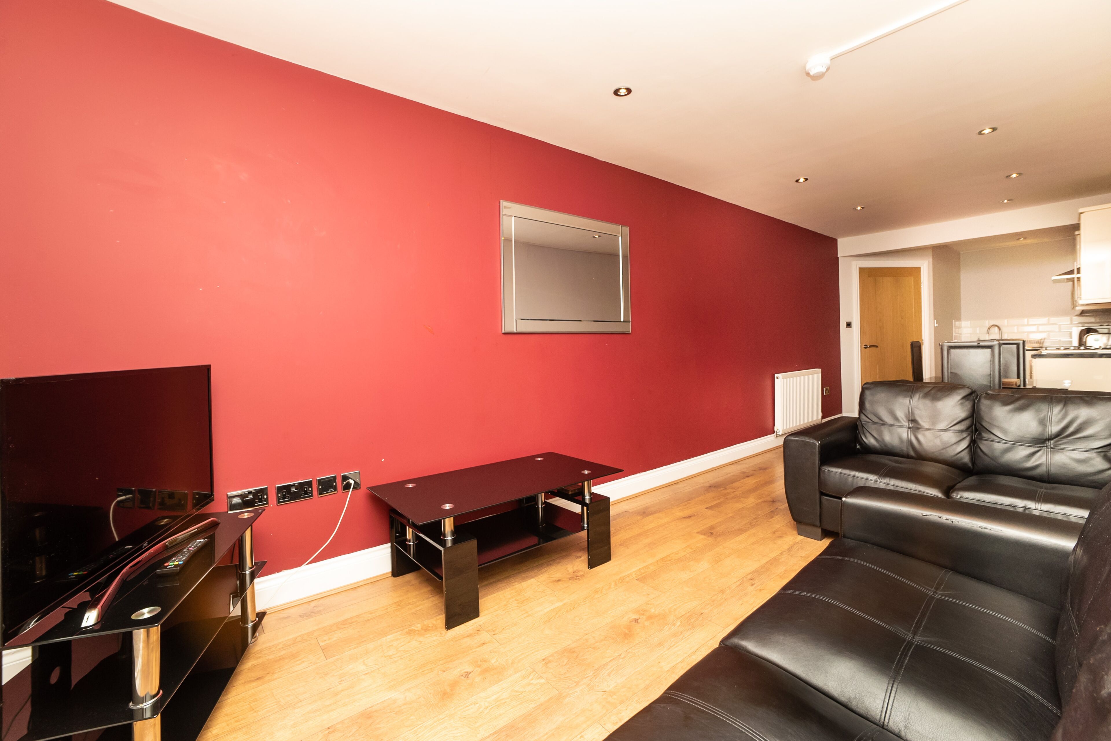A living room with a red wall, a black leather sofa, a wooden coffee table, and a flat-screen TV.