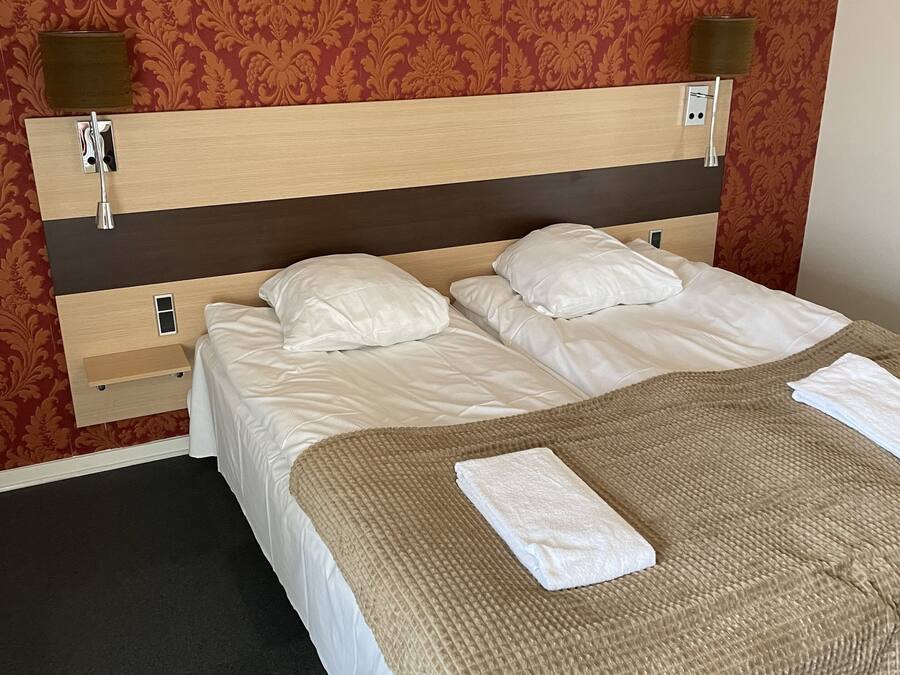 Un lit bien fait, avec du linge de lit blanc et une couverture marron, une tête de lit en bois à la teinte claire et un papier peint rouge à motifs.