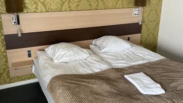 Una cama bien tendida con sábanas blancas, una manta de punto marrón y una toalla blanca. Hay dos lámparas de noche y un cabecero de madera.