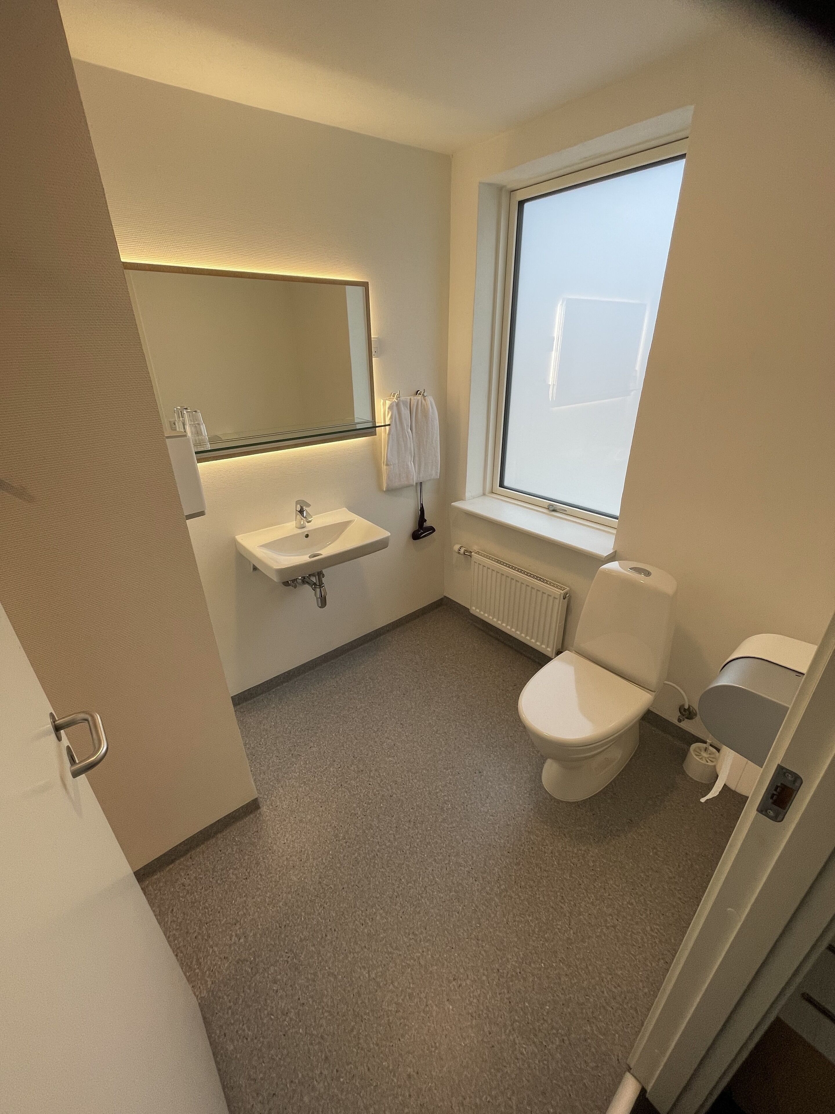 A small, modern bathroom with a white sink, mirror, and toilet. There is a window providing natural light and a towel rack with two towels.