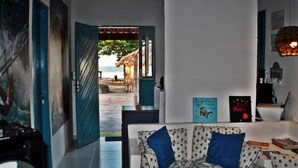 Ein Wohnzimmer mit einer Couch, einem Couchtisch und einem Bücherregal. Eine Tür führt auf einen Balkon mit Blick auf den Strand.