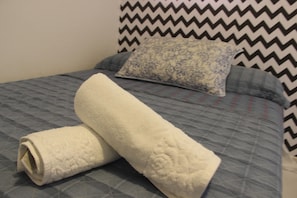 A bed with a blue quilted bedspread, a white rolled towel, and a patterned pillow. - El Surfista Hostel - Pousada (São Miguel do Gostoso)