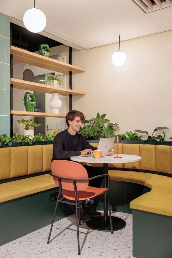 Una persona sentada a una mesa redonda con un ordenador portátil, rodeada por un sofá verde y sillas naranjas.