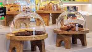 A bakery display with various baked goods under glass domes, a selection of packaged snacks, and condiments on shelves. - Maxhotel Amsterdam Airport Schiphol (Hoofddorp)