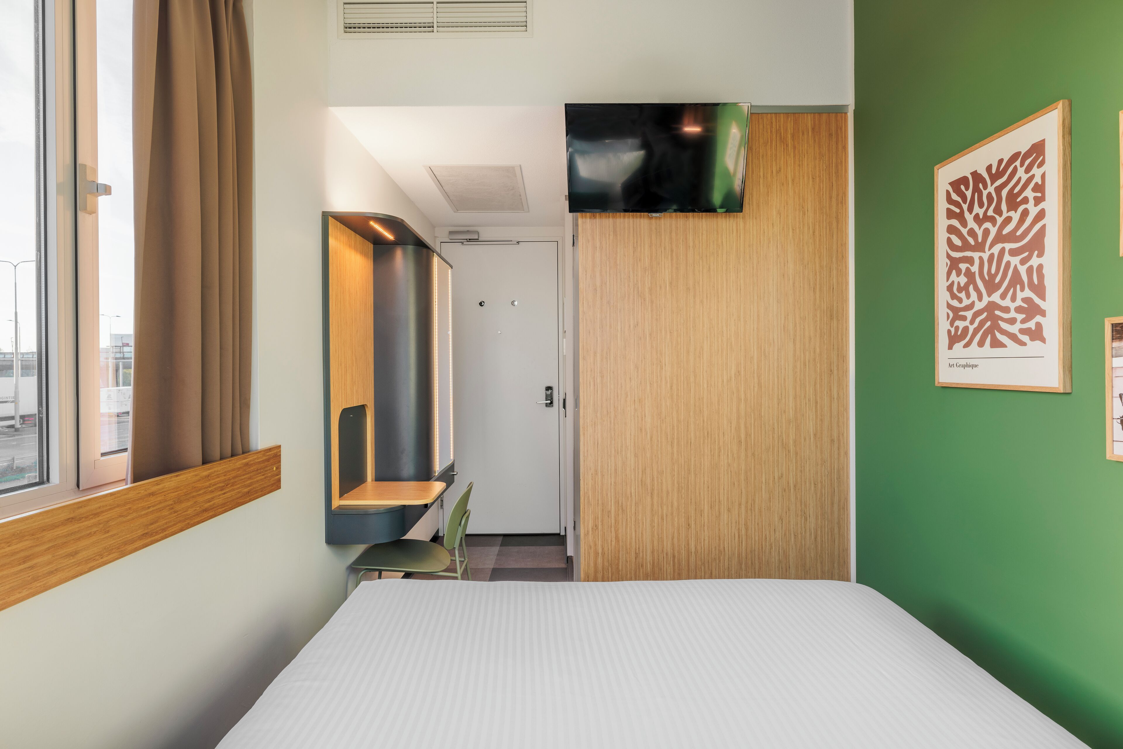 A hotel room with a bed, a desk, a chair, a TV, and artwork on the wall.