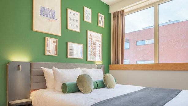 A neatly made bed with white linens and green pillows, a grey headboard, and framed artwork on the green wall. - Maxhotel Amsterdam Airport Schiphol (Hoofddorp)