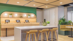 A modern reception area with a wooden counter, bar stools, and potted plants. - Maxhotel Amsterdam Airport Schiphol (Hoofddorp)