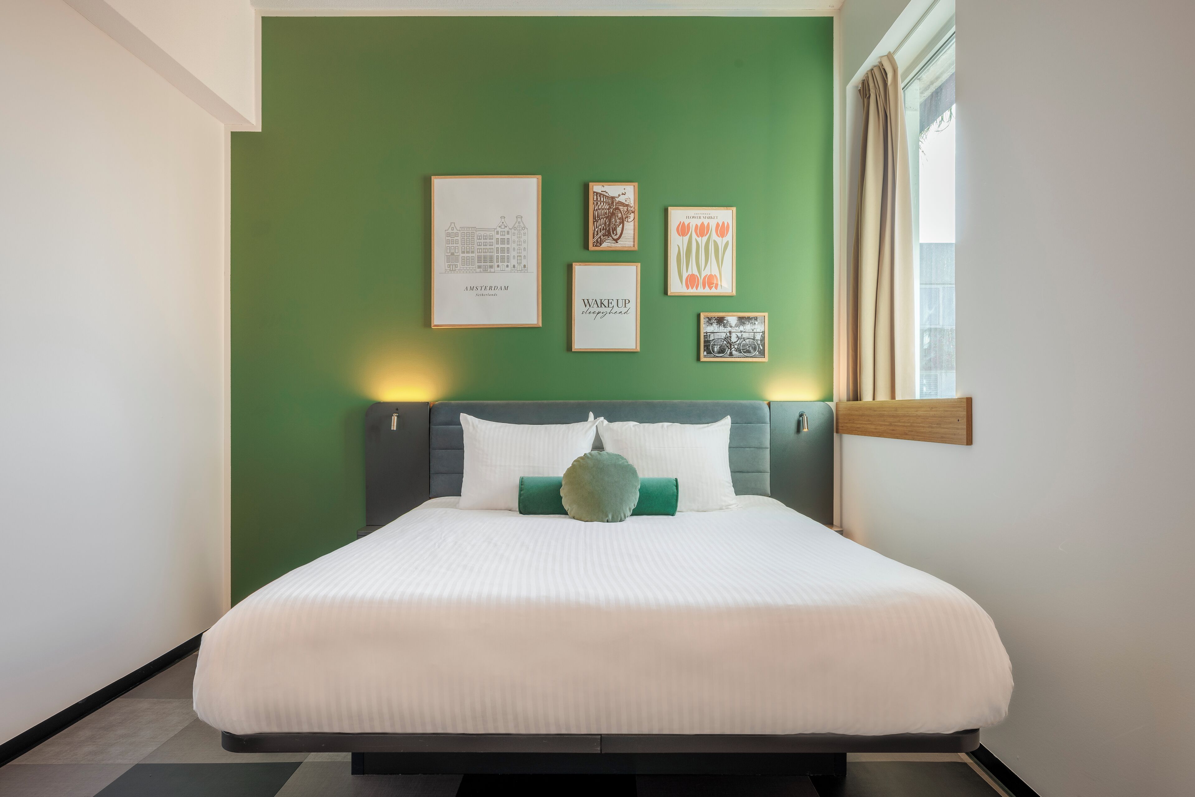 Un letto rifatto con lenzuola bianche, un cuscino verde e una testiera, con un muro verde sullo sfondo. In parete, sopra il letto, quattro quadri incorniciati.