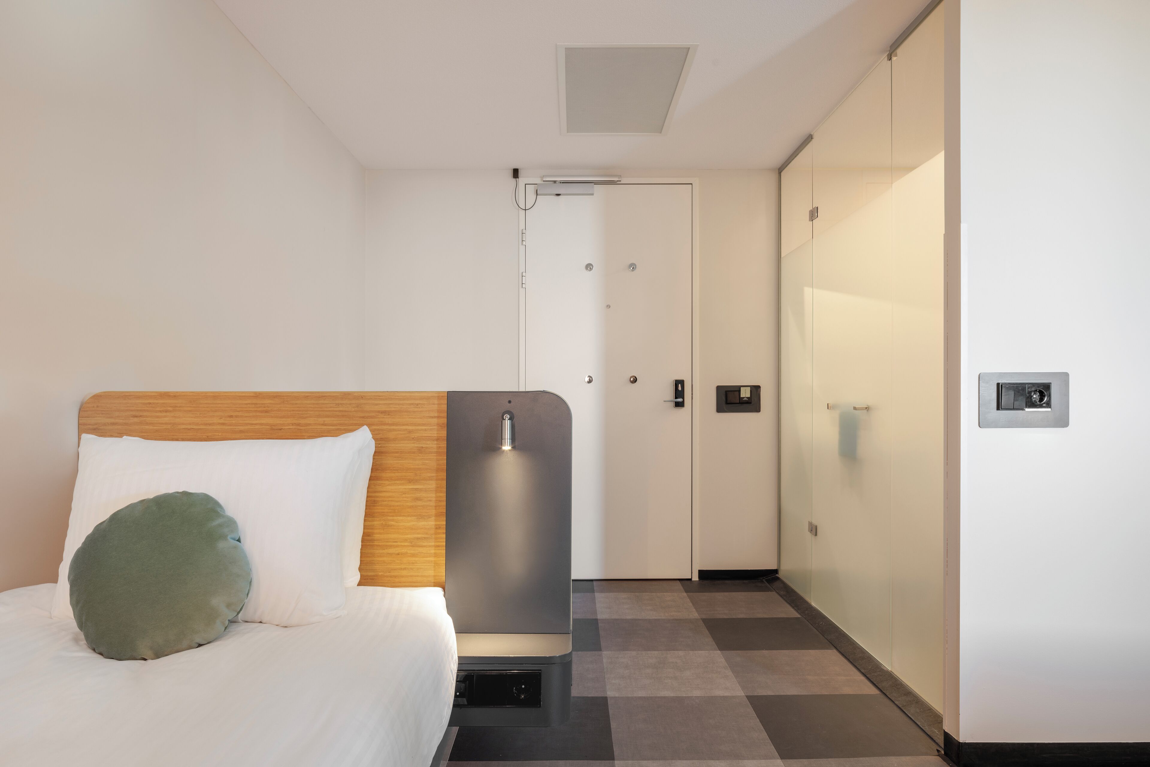 A hotel room with a bed, a bedside table, a wall-mounted light, a door, and a check-in kiosk.
