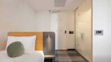 A hotel room with a bed, a bedside table, a wall-mounted light, a door, and a check-in kiosk.