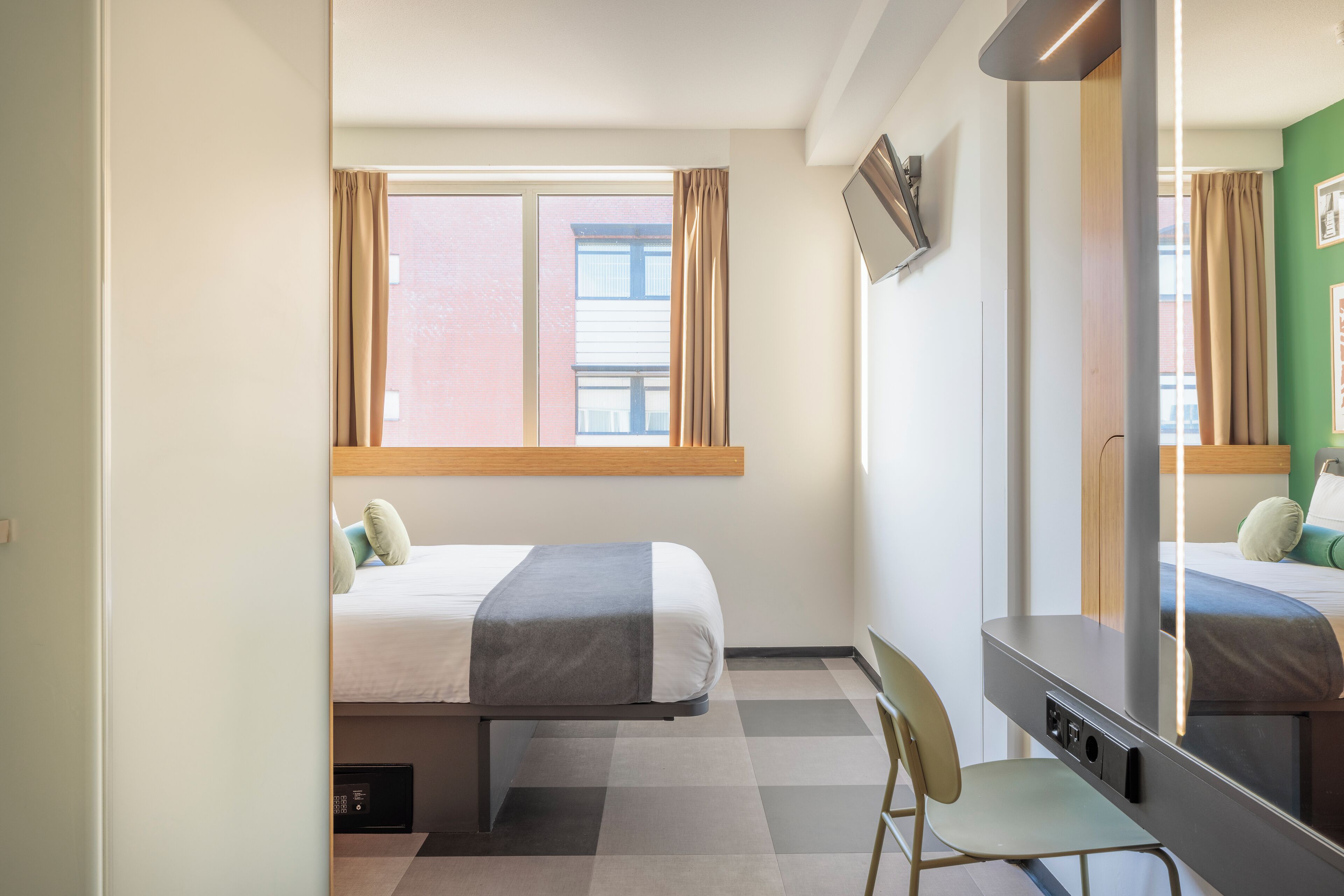 Una habitación de hotel con cama, escritorio, silla, espejo grande y ventana con cortinas.