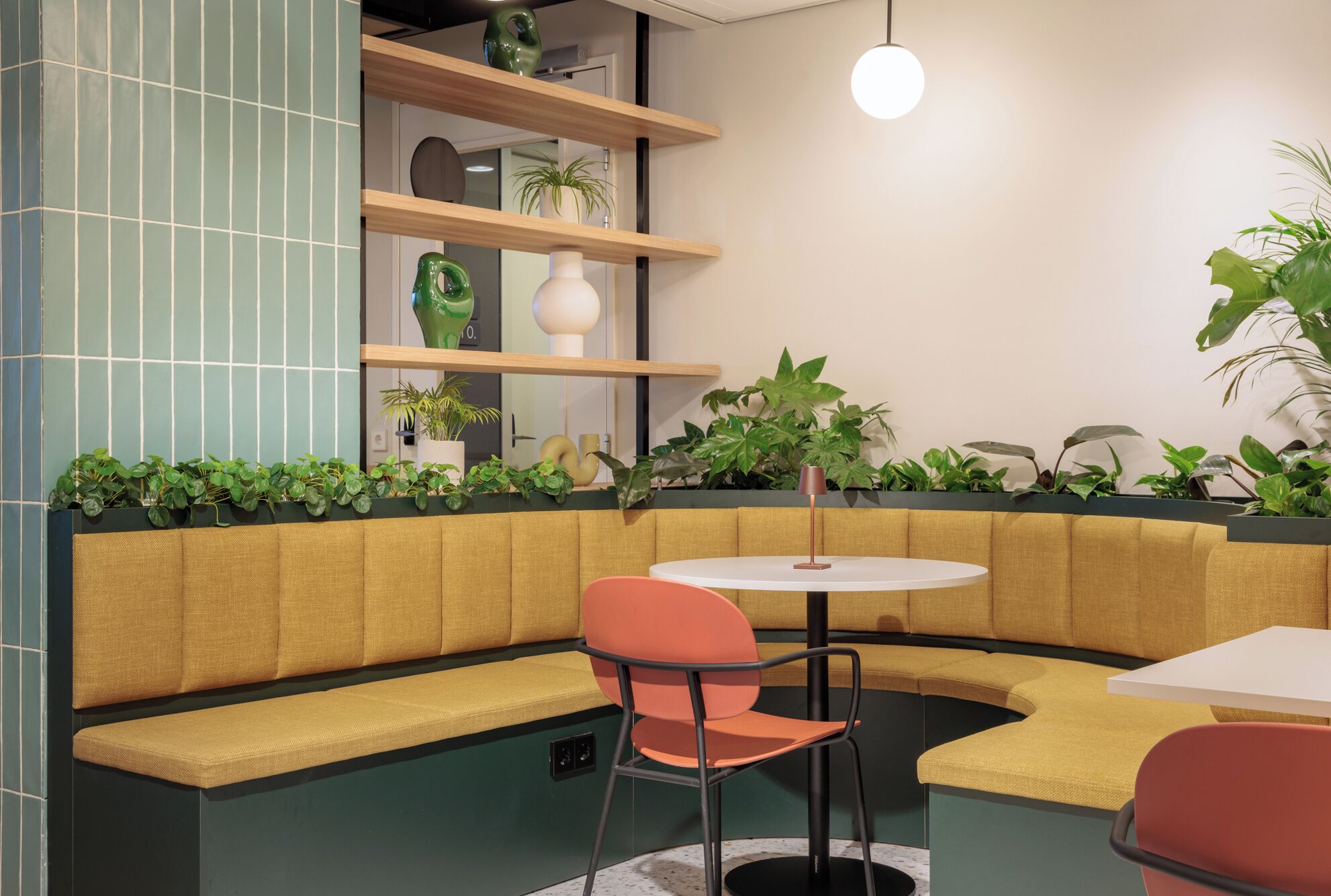 Un interior moderno con una zona de estar con banca curva, una mesa redonda y varias plantas.