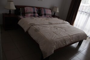 A neatly made bed with a quilt, two bedside lamps, and a window with curtains. - Gold Court Appartements Meublés (Yaoundé)