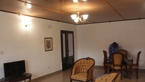 A living room with a television, a coffee table, and several chairs. There is a door and a chandelier. - Gold Court Appartements Meublés (Yaoundé)
