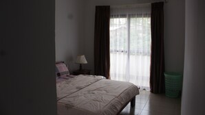 A bedroom with a bed, a nightstand, a lamp, a window with curtains, and an air conditioning unit. - Gold Court Appartements Meublés (Yaoundé)