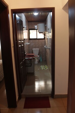 A narrow hallway leading to a bathroom with a toilet, shower, and sink. - Gold Court Appartements Meublés (Yaoundé)