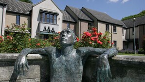 A sculpture of a reclining figure with outstretched arms, set against a backdrop of residential buildings and blooming flowers. - Brennan Court Guest Accommodation (Limerick)