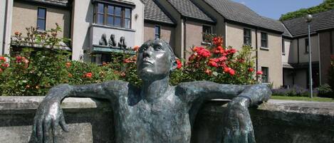 Une sculpture représentant une figure allongée, les bras tendus, se détachant sur un arrière-plan composé d’immeubles résidentiels et de fleurs en pleine floraison.