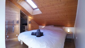 Un dormitorio con techo inclinado, paredes de madera, cama con ropa blanca, mesita de noche con lámpara y tragaluz.
